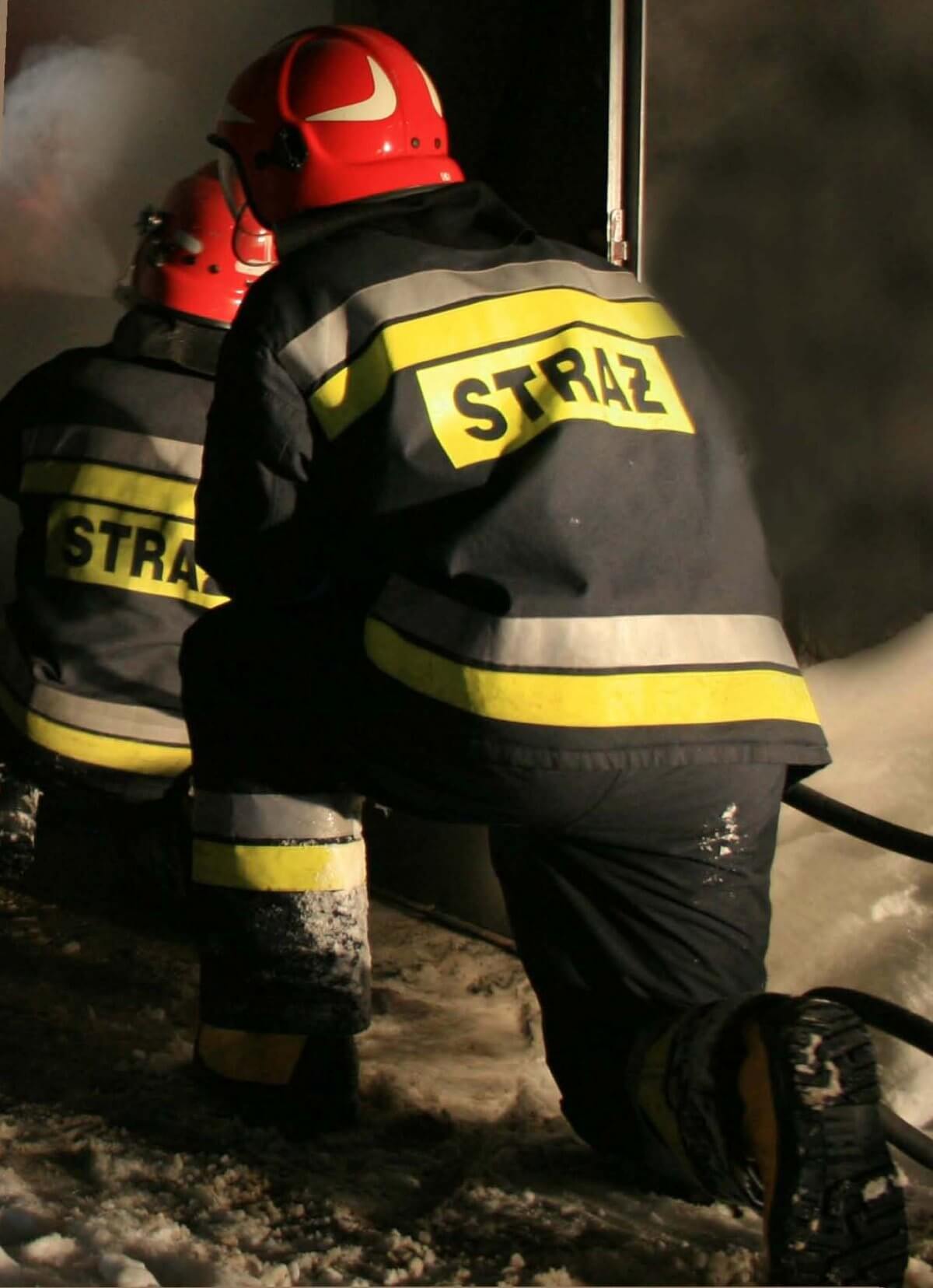 Strażak podczas akcji klęczący tyłem na prawej nodze, w czerwonym hełmie na głowie, w czarnym mundurze z napisem na plecach straż, a w tle za nim drugi strażak