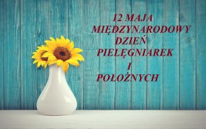 Na tle turkusowej ściany po prawej stronie napis 12 Maja Międzynarodowy Dzień Pielęgniarek i Położnych, po lewej w białym wazonie, na białym stole dwa żółte słoneczniki