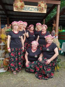 Panie z koła Gospodyń Wiejskich Mierzynianki na tle swojego stoiska podczas dożynek w Dobrej, ubrane w czarne koszulki i czarne spódnice w czerwone kwiaty, a na głowach kolorowe wianki