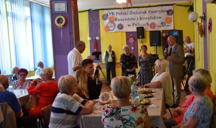 Siedziba Oddziału Rejonowego Polskiego Związku Emerytów,Rencistów i Inwalidów w Policach podczas uroczystości Dnia Inwalidy. Przemawia do mikrofonu Przewodniczący Rady Powiatu w Policach Cezary Arciszewski, obok stoją Panie z Zarządu Związku, a w tle sali przy stołach zasiadają przedstawiciele samorządu gminnego oraz Seniorzy.