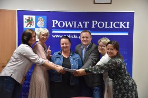 Podpisanie umowy na remont drogi powiatowej nr 3927Z Szczecin-Siadło Gróne, na tle granatowej grafiki z herbem Powiatu Polickiego po lewej i białym napisem Powiat Policki od lewej: przedstawiciel STRABAG, Małgorzata Schwarz Wójt Gminy Kołbaskowo, przedstawiciel STRABAG, Andrzej Bednarek Starosta Policki, Joanna Napiwodzka Wicestarosta Policki, Magdalena Sochanowska Naczelnik Wydziału Dróg Publicznych Starostwa Powiatowego w Policach