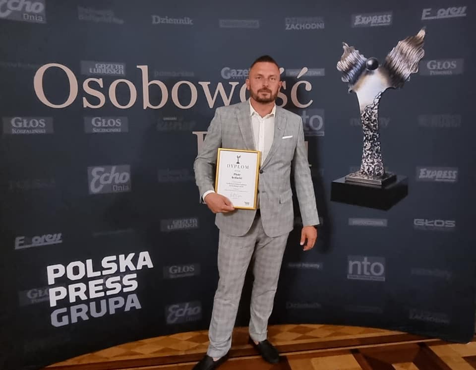 Pan Piotr Kołacki trzymający w prawej dłoni dyplom w ramce Tytuł Osobowości Roku Pomorza Zachodniego 2020, na tle ścianki w kolorze ciemnego granatu z napisami Osobowość, Polska Press Grupa i grafiką statuetki Osobowości Roku