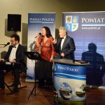 Występ artystyczny Teatru Rozmaitości GWITAJCIE, od lewej siedzący aktor grający na keybordzie, stojąca aktorka śpiewająca do mikrofonu, stojący aktor śpiewający do mikrofonu. W tle stojące grafiki Powiatu Polickiego z herbem na niebieskim tle.