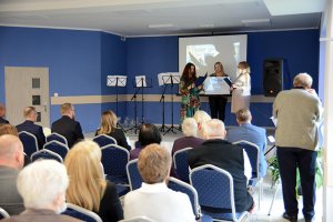 Naczelnik Wydziału Polityki Społecznej Halina Michalak i Prezes Stowarzyszenia Aleksandra Mazur-Woroniecka stoją na scenie przed siedzącymi ludźmi