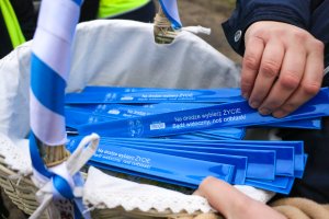 W koszyku wiklinowym, wyścielonym jasnym płotnem, w środku leżą samozaciskowe opaski odblaskowe w kolorze ciemnoniebieskim z grafiką od lewej strony herb Powiatu Polickiego z opisem obok, i biały napis Na drodze wybierz Życie. Bądź widoczny, nos odblaski.