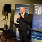 Przewodniczący Rady Powiatu w Policach Cezary Arciszewski stojący przy mównicy z grafiką Powiat Policki, witający wszystkich przybyłych na uroczystą premierę książki. w tle grafika Powiatu Polickiego z herbem na niebieskim tle.