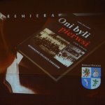 Slajd z białym napisem w lewym górnym rogu PREMIERA, poniżej po środku okładka czwartej części książki Oni byli pierwsi autorstwa Stefana Marszałka, w prawym dolnym rogu herb Powiatu Polickiego