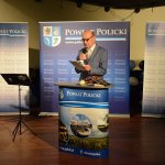 autor książki Oni byli pierwsi Stefan Marszałek stojący przy mównicy z grafiką Powiat Policki, czytający z kartki słowo. w tle grafika Powiatu Polickiego z herbem na niebieskim tle.