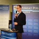 Bartosz Sitarz stojący przy mównicy z grafiką Powiat Policki, wygłaszający swoją prelekcję. w tle grafika Powiatu Polickiego z herbem na niebieskim tle.