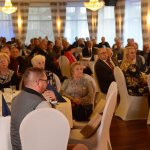 Zbliżenie gości, siedzących przy okrągłych stołach i słuchających wystąpień.