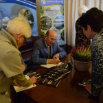 Stefan Marszałek siedziący na skórzanej sofie przy stoliku, podpisujący swoją książkę. Na stoliku lezą rozłożone egzemplarze publikacji, a na środku szara doniczka z kolorowym wrzosem. W tle grafika Powiatu Polickiego z herbem,