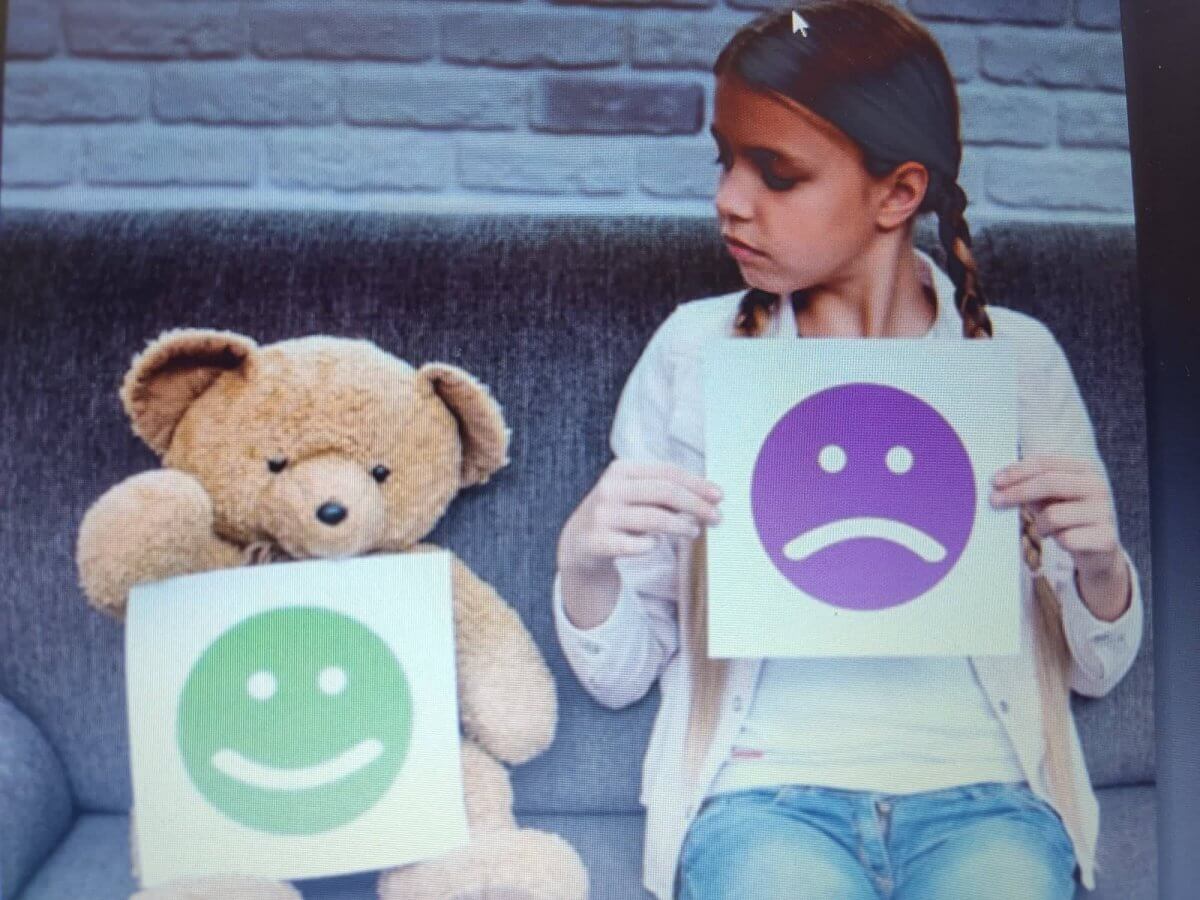 Na tapczanie siedzi miś trzymający emotkę z uśmiechem, obok niego dziewczynka trzymająca emotkę ze smutną miną