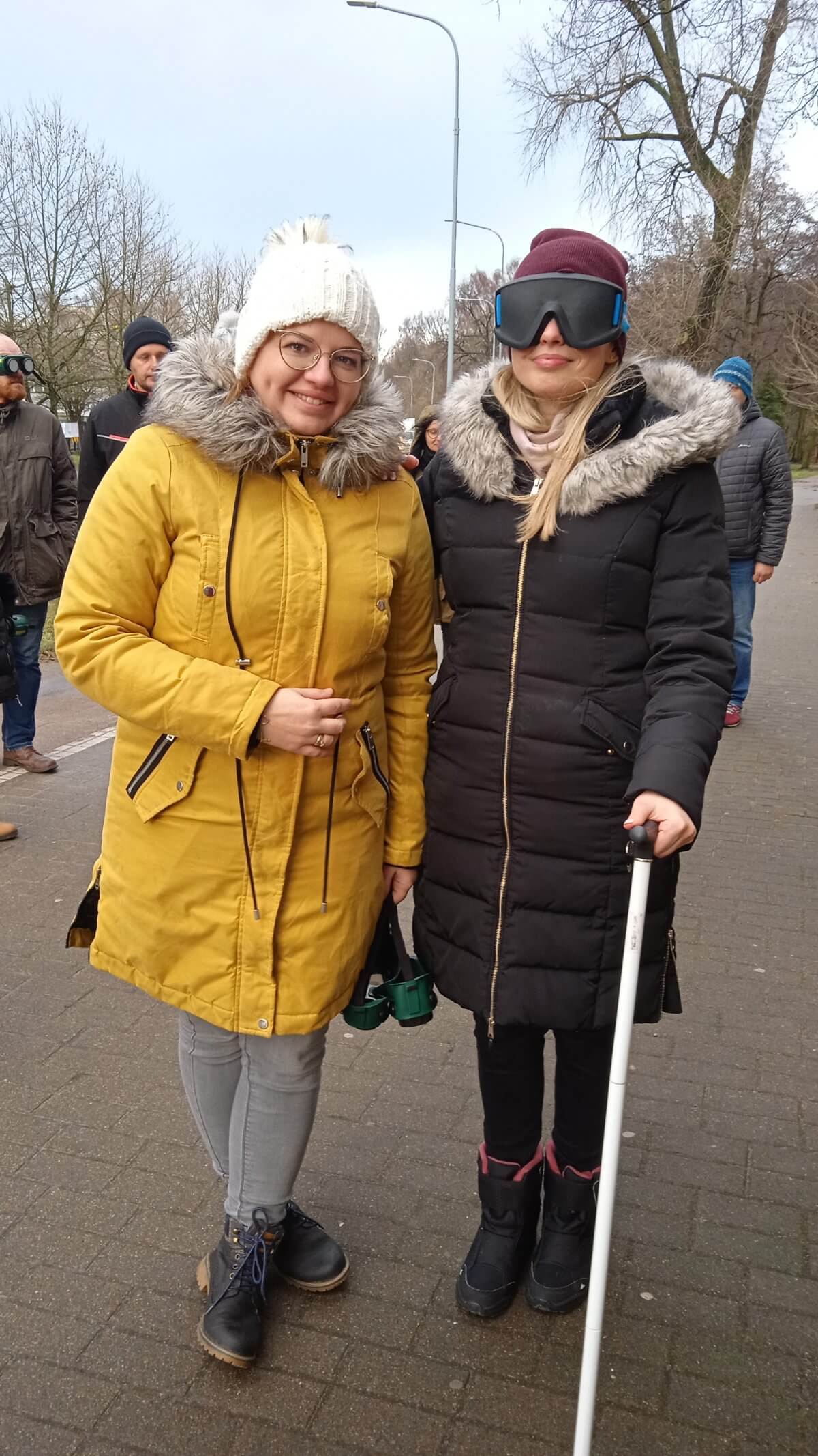 Dwie osoby stoją, w tym jedna z laską i goglami