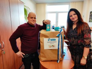 Prezes Fundacji Polskich Wartości Wojciech Woźniak trzymający w lewej dłoni czekoladę z grafiką zajączka, oparty o karton opakowany szarym papierem, z naklejoną kartką z herbem Powiatu Polickiego u góry i pod nim napisem RODAKOM NA KRESACH Wielkanoc 2022, a po prawej stronie oparta o ten sam karton Naczelnik Wydziału Informacji i Promocji Agnieszka Rychlik-Bakalarz