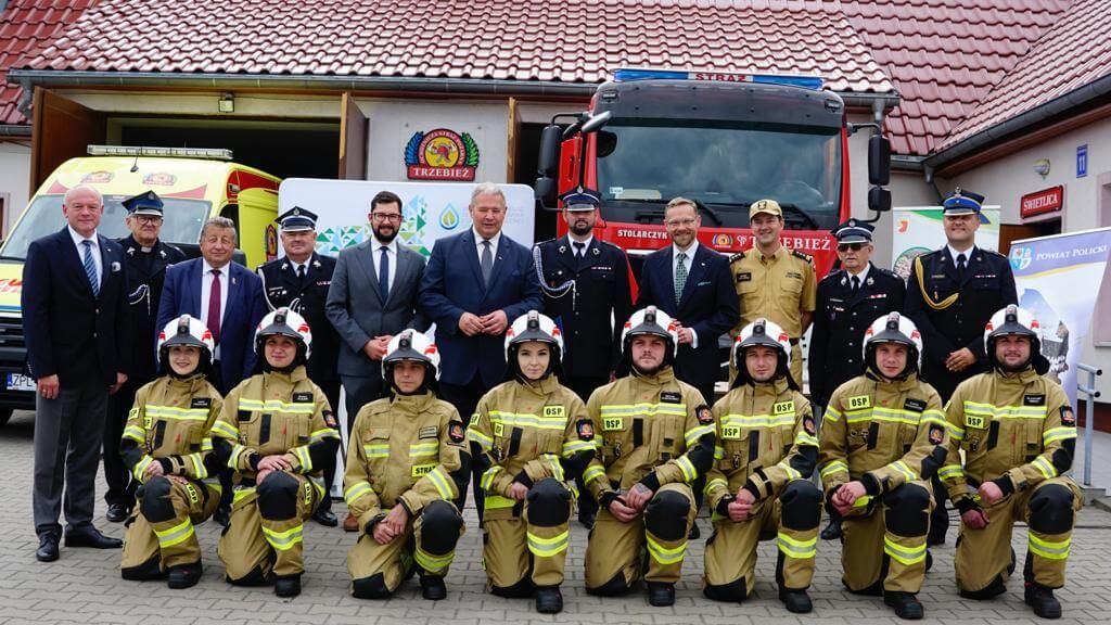 Kilkunastoosobowa grupa ludzi pozujaca do zdjęcia na tle wozu strażackiego.W pierwszym rzędzie w żółtych mundurach strażacy.