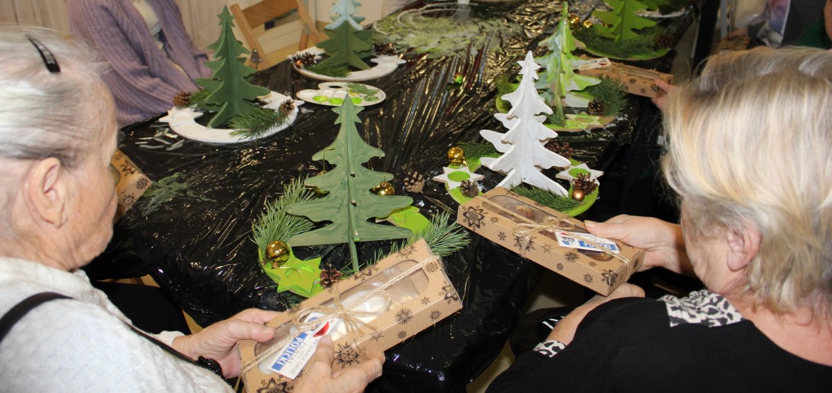 Seniorki siedzą przy stole z opakowaniami pierniczków od Starostwa Powiatowego w Policach, na stole znajdują się zielone choinki