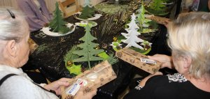 Seniorki siedzą przy stole z opakowaniami pierniczków od Starostwa Powiatowego w Policach, na stole znajdują się zielone choinki