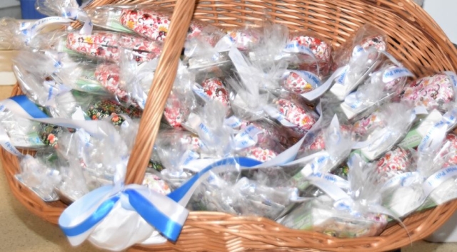 W koszu wiklinowym ułożone upominki zapakowane w celafon zawiązane w białą wstążkę z napisem w kolorze niebieskim POWIAT POLICKI