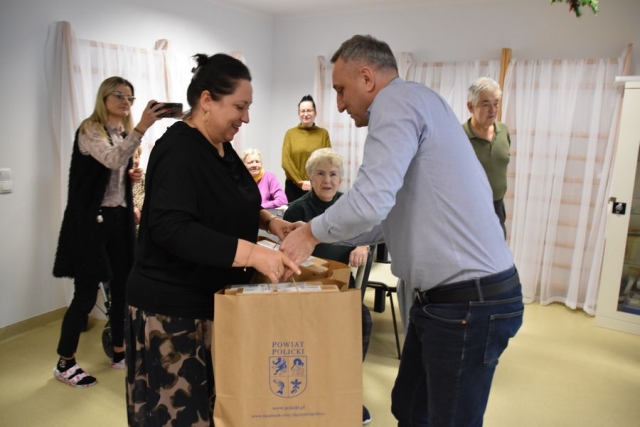 Przedstawiciel Powiatu Polickiego wręcza do rąk Kierownikowi Dziennego Domu Senior Plus dwie duże torby papierowe z laptopami i tabletami, a w tle siedzący przy stole uczestnicy placówki