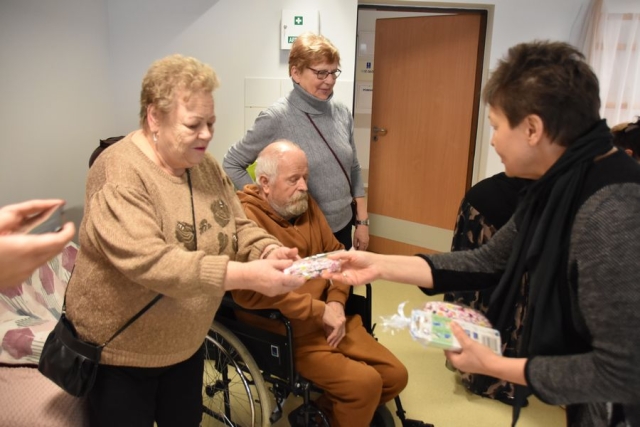 Członek Zarządu Powiatu w Policach Beata Chmielewska wręcza uczestniczce Dziennego Domu Seniora Plus, stojącej obok mężczyzny siedzącego na wózku, upominek