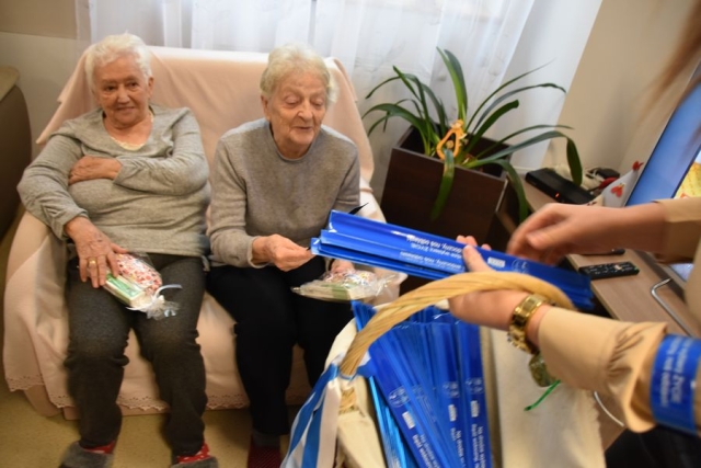 Na sofie siedzą dwie kobiety, którym wręczane zostają opaski odblaskowe w kolorze granatowym z wiklinowego koszyka