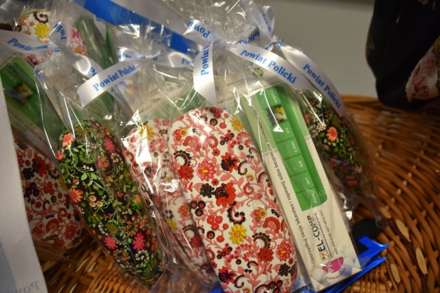 W koszu wiklinowym ułożone upominki zapakowane w celafon zawiązane w białą wstążkę z napisem w kolorze niebieskim POWIAT POLICKI