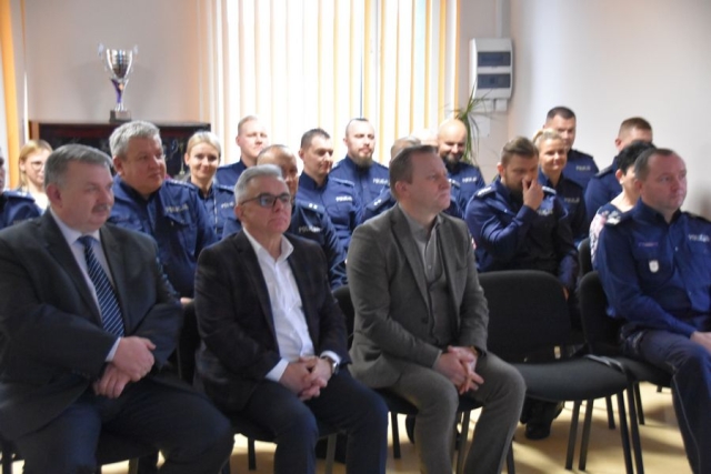 Uczestnicy odprawy policyjnej siedzący na krzesłach ustawionych w rzędach.