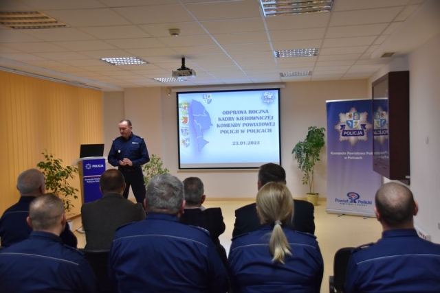 Sala odprawy policyjnej, siedzący na krzesłach uczestnicy odprawy, przed nimi na ekranie slaid prezentacji i Komendant Wojewódzki Policji