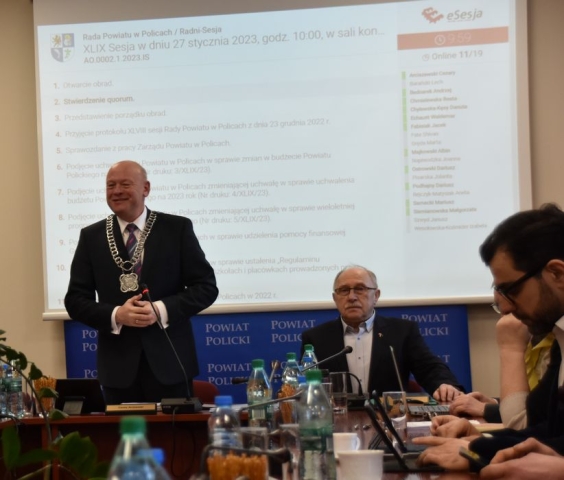 Obrady sesji Rady Powiatu w Policach, przy stołach ustawionych w literę U zasiadają Radni Powiatu Polickiego, przy stole prezydelialnym wszystkich przybył na sesję wita na stojąco Przewodniczący Rady Powiatu na tle ekranu wyświetlającego z projektora porządku obrad sesji