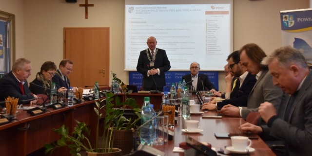 Obrady sesji Rady Powiatu w Policach, przy stołach ustawionych w literę U zasiadają Radni Powiatu Polickiego, przy stole prezydelialnym wszystkich przybył na sesję wita na stojąco Przewodniczący Rady Powiatu na tle ekranu wyświetlającego z projektora porządku obrad sesji