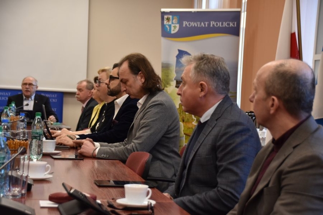 Obrady sesji Rady Powiatu w Policach, przy stole zasiadają radni Powiatu Polickiego w tle stoi grafika z herbem i napisem Powiat Policki
