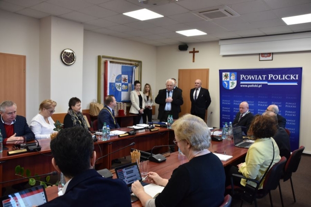 Obrady sesji Rady Powiatu w Policach, przy stołach zasiadają radni Powiatu Polickiego, słuchają przemówienia stojącego męźczyzny z mikrofonem-Przewodniczącego Okręgowej Rady,za nim stoją dwie kobiety i dwóch mężczyzn