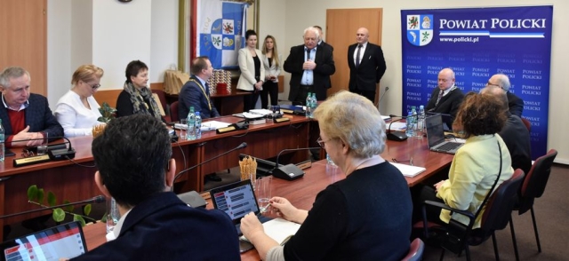 Obrady sesji Rady Powiatu w Policach, przy stołach zasiadają radni Powiatu Polickiego, słuchają przemówienia stojącego męźczyzny z mikrofonem-Przewodniczącego Okręgowej Rady,za nim stoją dwie kobiety i dwóch mężczyzn