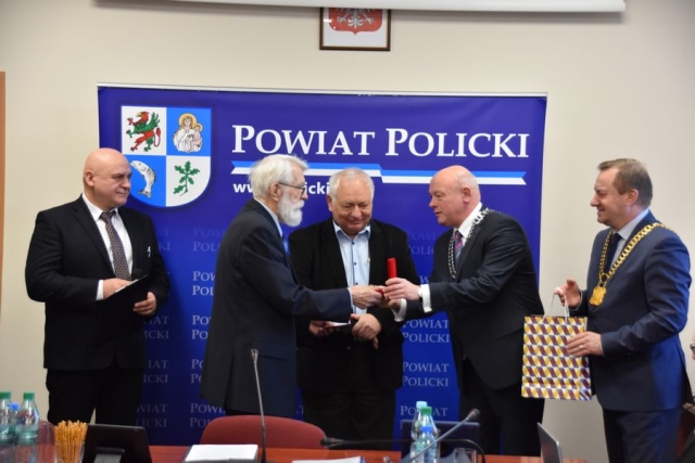 Obrady sesji Rady Powiatu w Policach, na tle grafiki niebieskiej z dużym herbem Powiatu Polickiego stoi pięciu mężczyzn, następuje wręczenie medalu i gratulacji od Przewodniczącego Rady Powiatu w Policach i Starosty Polickiego