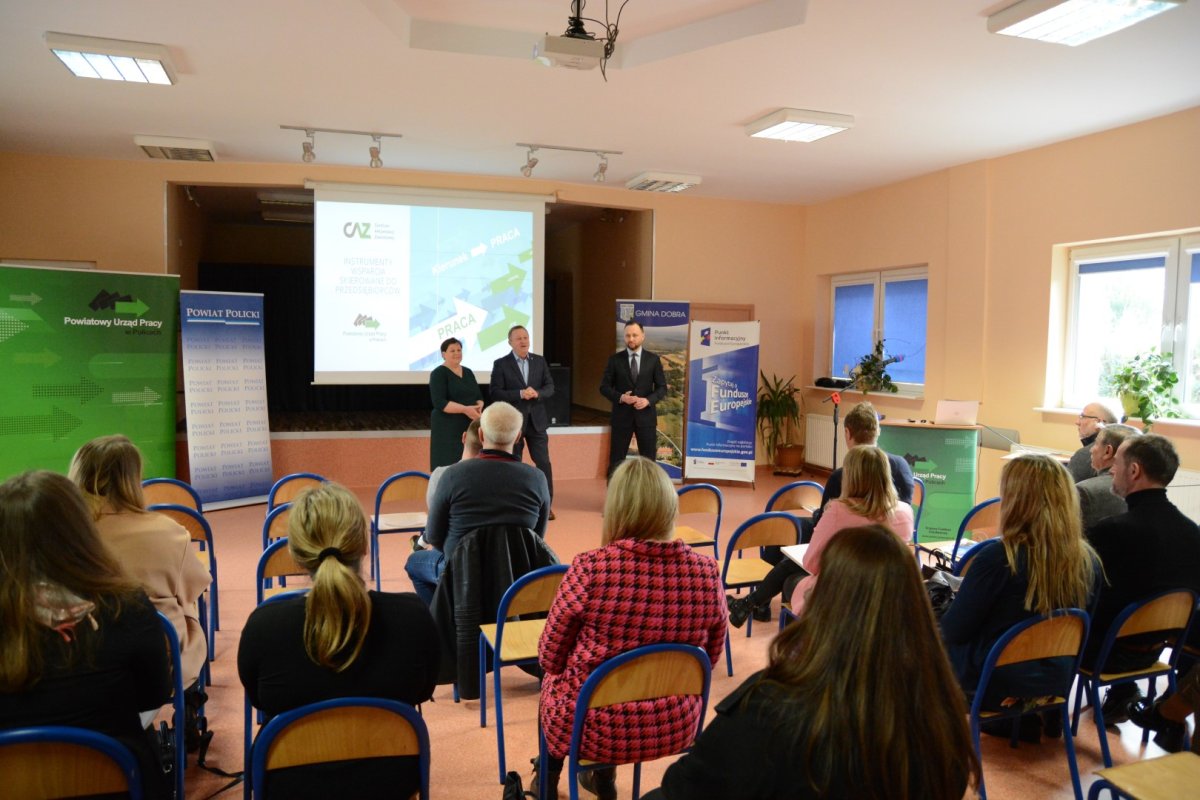 Spotkanie informacyjne, siedzący ludzie tyłem, a w tle 3 osoby stoją