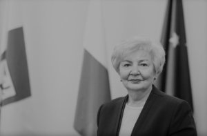 Czarno-białe zdjęcie, na tle dwóch flag polskiej i Unii Europejskiej stoi kobieta patrząca się na wprost