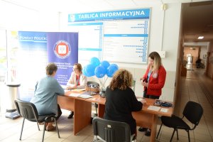 Stoisko sanepidu w urzędzie, a przy stoisku trzy osoby siedzące i jedna stojąca