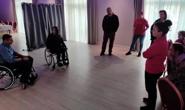 Na sali szkoleniowej, przy stołach przykrytych białymi obrusami siedzące kobiety, a przed nimi na wózkach siedząca kobieta i mężczyzna oraz stojący uczestnicy szkolenia