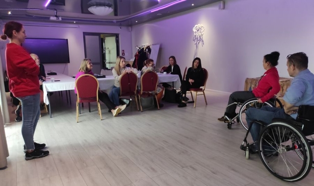 Sala szkoleniowa, przy stołach przykrytych białymi obrusami siedzi pięć kobiet, a przed nimi na wózkach siedząca kobieta i mężczyzna