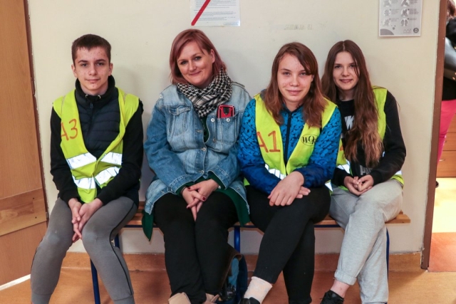 Na ławce siedzi chłopak w odblaskowej kamizelce wordu, uśmiechajaca się dorosła kobieta w jasnych włosach oraz dwie młode dziewczynki w kamizelkach odblaskowych word
