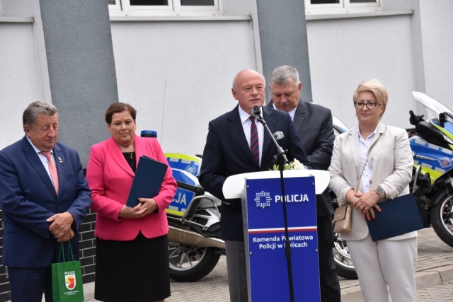 Cezary Arciszewski Przewodniczący Rady Powiatu w Policach w trakcie wspólnego składania życzeń funkcjonariuszom policji wraz z Joanna Napiwodzką Wicestarostą Polickim, Władysławem Diakunem Burmistrzem Polic, Teresą Derą Wójtem Gminy Dobra, Jarosławem Burbą Burmistrzem Nowego Warpna na dziedzińcu komendy w tle żółto-niebieskiem motocykle policyjne.