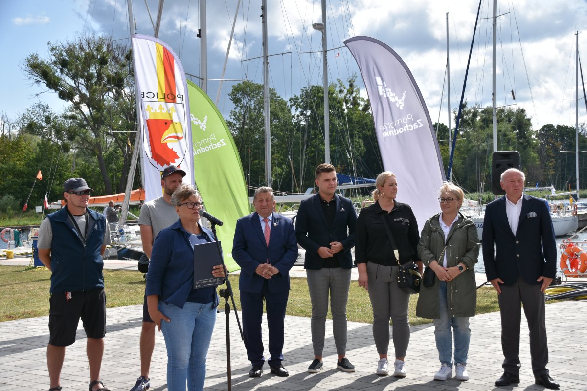 Grupa mieszana, mężczyźni, kobiety, przedstawiciele Powiatu Polickiego i Gminy Police w trakcie oficjalnego otwarcia regat.Przy mikrofonie przemawia przedstawicielka Zachodniopomorskiego Oddziału Związku Zeglarskiego, w tle flagi sponsorów i żaglówki zacumowane w porcie. Wietrzny, słoneczny dzień.