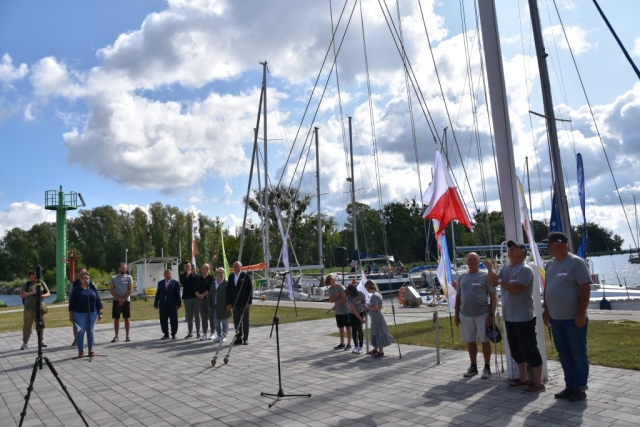 Moment wciągniecia biało-czerwonej flagi na maszt.W tle uczestnicy wydarzenia oraz zacumowane żagłówki w porcie.