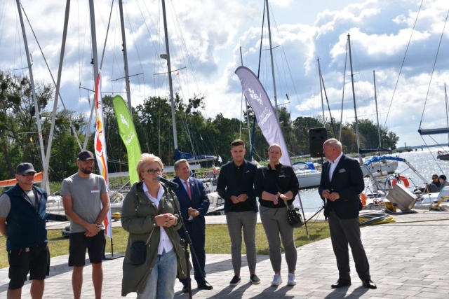 Moment przemówienia Joanny Napiwodzkiej Wicestarosty Polickiego podczas otwarcia regat.W tle pozostali przedstawiciele samorządów i organizatorów oraz powiewające na wietrze flagi sponsorów i zacumowane żaglówki w porcie.
