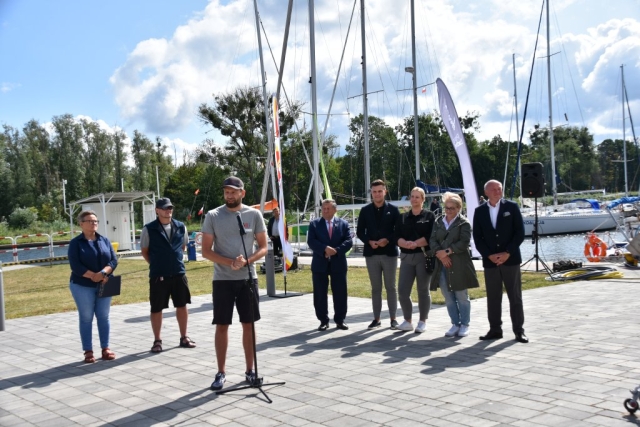 Moment wypowiedzi przedstawiciela Zachodniopomorskiego Związku Żeglarskiego, W tle grupa mieszana mężczyźni, kobiety, przedstawiciele samorzadów oraz zacumowane w porcie żaglówki.