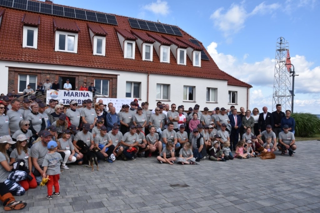 Kilkudziesięcioosobowa grupa ludzi, mężczyźni, kobiety pozująca do zdjęcia grupowego na tle białego budynku z czerwonym dachem.