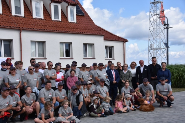 Kilkudziesięcioosobowa grupa ludzi, mężczyźni, kobiety pozująca do zdjęcia grupowego na tle białego budynku z czerwonym dachem. Uczestnicy, organizatorzy i sponsorzy regat.