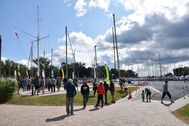 Tłum ludzi, męzczyźni, kobiety oczekujący na oficjalne otwaercie regat w Marinie w Trzebieży. W tle żaglówki zacumowane w porcie. Słoneczny, wietrzny dzień.