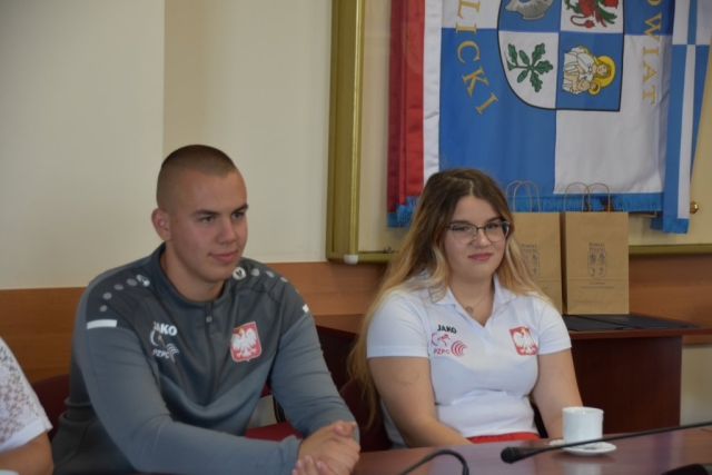 Siedząca przy stole zawodniczka, kobieta w długich jasnych włosach i okularach oraz mężczyzna, w którtkich ciemnych włosach, obie osoby mają na piersi oznakowanie z białym orłem w koronie na czerwonym tle oraz z logiem Polskiego Związku Podnoszenia Ciężarów przedstawiającym gryfa.