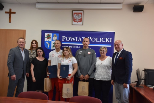 Zdjęcie grupowe, mężczyźni i kobiety, Przedstawiciele Powiatu Polickiego oraz dwoje sportowców, w tym mama zawodniczki oraz trener na tle ścianki z herbem Powiatu Polickiego.